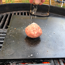 Load image into Gallery viewer, A smash burger being seasoned on a plancha with an Axel Perkins Heirloom Burger Press in the background 
